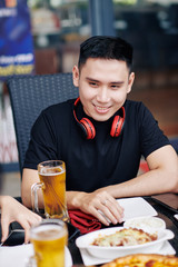 Happy Asian young man sitting at the table with food and alcoholic drinks and smiling while talking to his friends in cafe