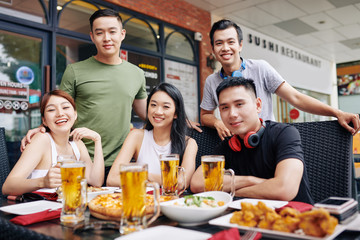 Portrait of Asian young friends smiling at camera while sitting at the table with glasses of beer during their meeting at the restaurant