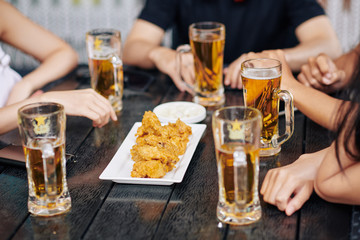 Close-up of friends sitting at wooden table and drinking beer from the glasses and eating fried chicken in cafe
