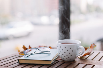 Close up photo of glasses, white mug with tea, green book and orange maple leaves on wooden table. Comfort, coziness and hygge concept. Autumn mood. Thanksgiving day. Still life. Selective focus.