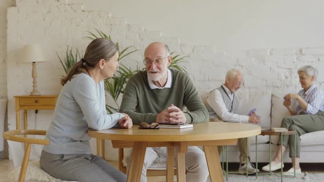Tracking shot of senior man and woman sitting at table and talking joyfully, another elderly couple playing cards in background sitting on sofa in common room of nursing home