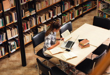 Tired young female student study in the library reading a book and using laptop for researching online.