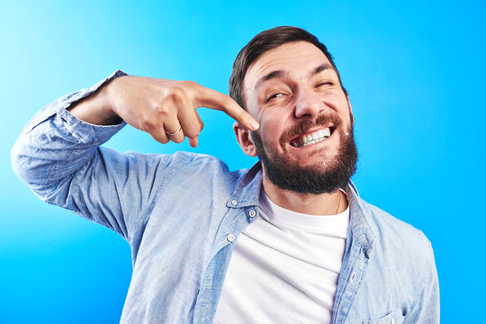 A young Caucasian man shows a finger in his mouth pointing to white teeth and a wonderful smile. Funny and funny. Dental health. On in the studio on a blue background