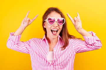 Yeah, feeling cool and awesome. Excited happy funny young  woman lifts mood from loud music, shows rock n roll gesture, in glasses in the shape of a heart on a yellow bright background