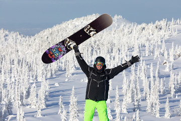 snowboarder in a black jacket and helmet, in bright green pants standing with snowboard raised...