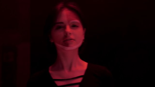 pretty brunette girl with paint on face in black dress poses at bright light rays in dark red room closeup slow motion
