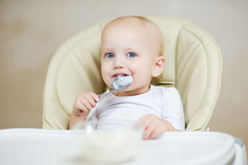 cute baby girl in a feeding chair holds a spoon in his mouth and smiles