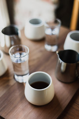 White cups of black coffee, steel coffee pots, glasses of water, wooden table