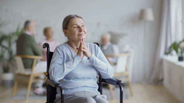 Tracking shot of disabled senior woman in wheelchair looking away and contemplating in nursing home, other aged patients in background