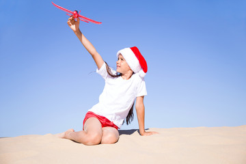 Christmas celebration vacations on a tropical beach: Happy cute little girl wearing a red Santa hat sitting on the sandy beach by the sea and playing with the red toy plane on clear sunny day