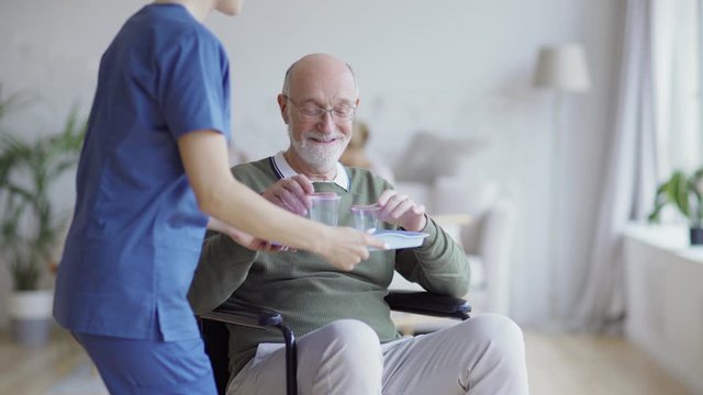 Disabled elderly man in eyeglasses sitting in wheelchair and looking away thoughtfully. Senior patient smiling and looking at camera when female nurse bringing him vitamin pills and glass of water