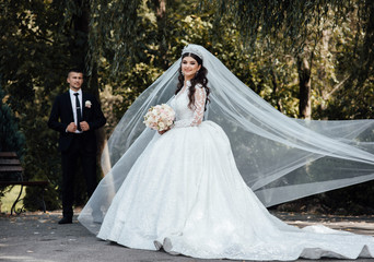 Fototapeta premium Wedding couple. Beautiful bride and groom. The groom kisses the bride's hand. Woman in fashionable outfit waving veil wind. Stylish wedding couple walking in love