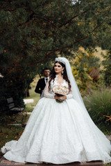 Handsome groom in black formal suit and his elegant bride in dress and veil with bouquet with beautiful hairdress. Pair embrace on a walk in the big green park. Beautiful wedding photosession.