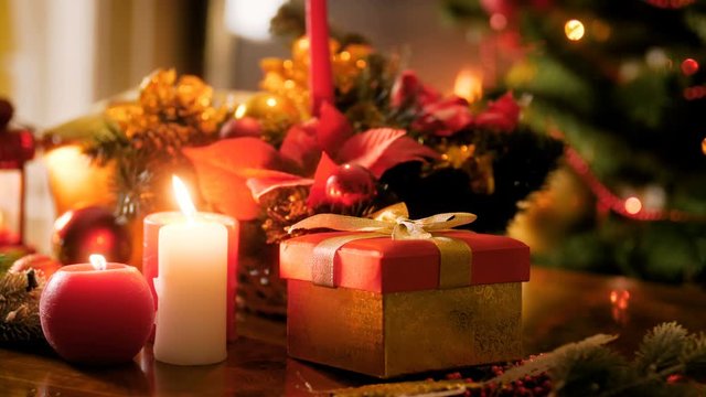 Closeup 4k video of Christmas gift and burning candle on wooden table against glowing Christmas lights and garlands. Perfect shot for winter celebrations and holidays