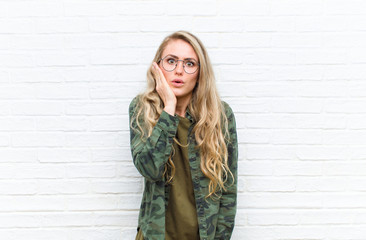 young blonde woman feeling shocked and astonished holding face to hand in disbelief with mouth wide...