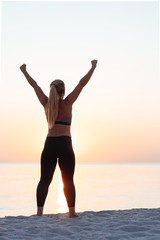 Young sporty woman standing by the sea with her arms raised in the air in sunset