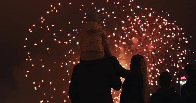 Happy parents with a small child look at the sky and enjoy the party. Silhouette of people watching explosive and colorful fireworks in the evening sky. Holiday event and celebration. People have fun.