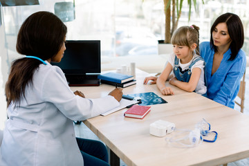 Obraz premium Side view of African-American female doctor discussing over x-ray report with Caucasian mother and daughter in the modern office at hospital. Laptop, nebulizer, x-ray on the table