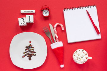 New year composition Calendar December 31st. Sweet chocolate Christmas tree on plate, cutlery in santa hat, cup of cocoa with marshmallows, blank notepad on red background. Flat lay Mockup Template
