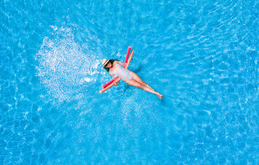 Zenith aerial view of a swimming pool in summer. Young girl in a swimsuit and hat floating with...