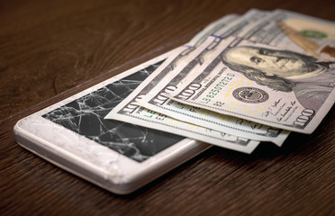 One hundred dollar bills lie on a broken smartphone. Phone repair fee._
