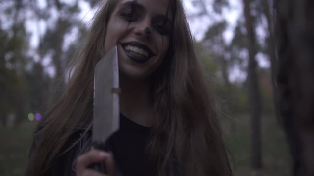 Crazy young woman with Halloween clown makeup on her face holding big knife in hand, laughing. The girl looking angrily in the camera and hits the trunk of the tree with a cleaver