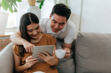 young asian love couple drinking coffee and using mobile tablet, watching movie online on sofa at home office, internet technology, social media, online booking, entertainment, shopping online concept