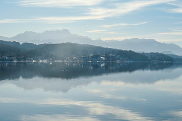 A view on a lake and Alps in the back. The calm surface of the lake is reflecting the mountains, sunbeams and clouds. Little city located at the lake side. Church with bell tower in the middle