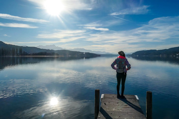Fototapeta na wymiar Girl wearing a jacket and beanie stands at the end of a promenade on the lake. Soft reflections of the clouds in the lake. Clear but cloudy day. High mountains in the back Calmness and relaxation