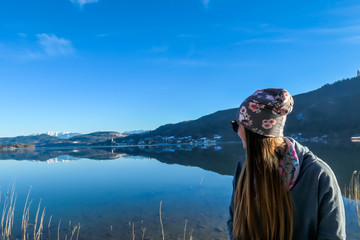 Fototapeta premium Girl wearing a beanie poses with the lake view behind her. Calm surface of the lake reflects the mountains and the sky. Girl is smiling, enjoying her time and beautiful weather. Happy autumn moment.