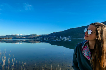 Girl wearing a beanie poses with the lake view behind her. Calm surface of the lake reflects the mountains and the sky. Girl is smiling, enjoying her time and beautiful weather. Happy autumn moment.