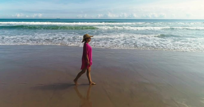 bella mujer con vestidos rosa y pamela pasea relajada por la playa y un dron le sigue