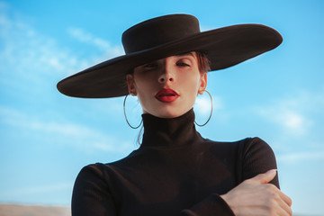 Close up portrait of a sexy beautiful mysterious woman with red lips and big black hat on. Stunning woman posing sensually outdoors on sunset under blue sky