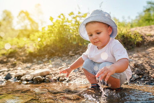 Portrait of playful small little boy child playing with rocks and water by the lake or river in sunny day wearing hat in nature