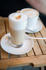 Glas Latte Macchiato und Tasse Cappuccino Kaffee mit Milchschaum auf Holztisch draußen im Straßencafé 