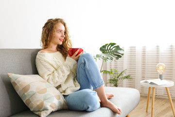 Young beautiful woman wearing white sweater on grey textile sofa at home. Attractive slim female in...