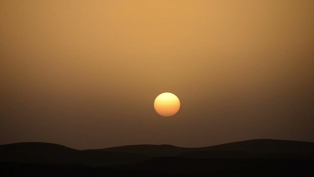 A big yellow sun setting in the front of a golden sky in the Sahara desert in Morocco, with dark dunes in the horizon. Timelapse. 1800% accelerated