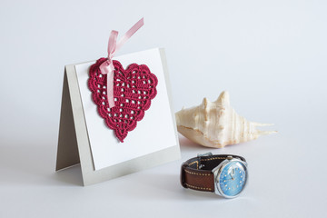 Greeting card with knitted heart. Hearts are crocheted with  red threads. Holiday decor. Still life with greeting card, watch and sea shell 