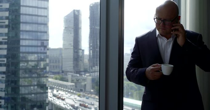 A brutal bald man in glasses and a business suit is standing at the large window of a modern office center with coffee and talking on the phone