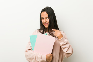 Young hispanic woman holding some notebooks pointing with finger at you as if inviting come closer.