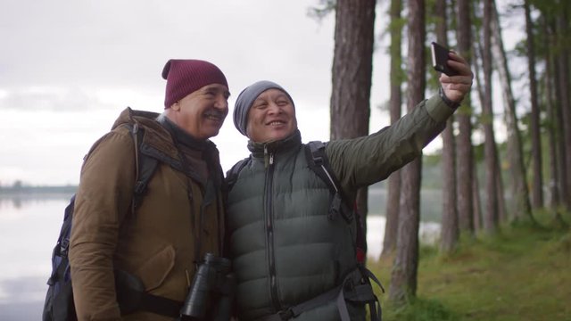Two joyous senior men smiling at smartphone camera and taking selfie against beautiful lake while having autumn hiking trip