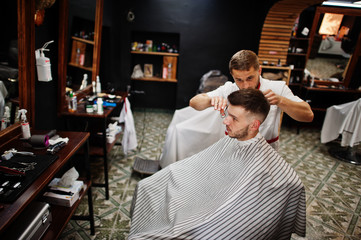 Young bearded man getting haircut by hairdresser while sitting in chair at barbershop. Barber soul.