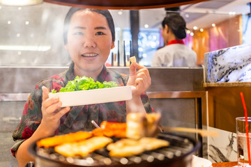 Happy Asian woman holding a tray of green vegetables and garlic with a stove of grilled meat in front side