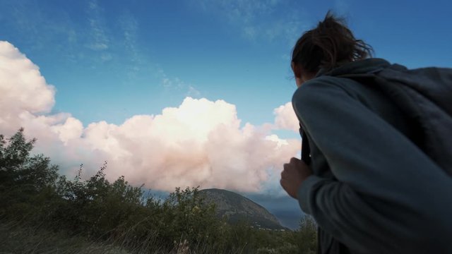 woman hiker looking beautiful colorful amazing sunset sky. female hiker with backpack standind on mountain slope at seaside overlooking of amazing big rose clouds on blue sky over seaside at sunset