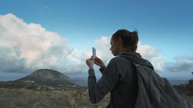 woman hiker looking amazing beautiful cloudy sunset sky and taking photo using mobile phone. female hiker with backpack standind on mountain slope at seaside overlooking of amazing big rose clouds on