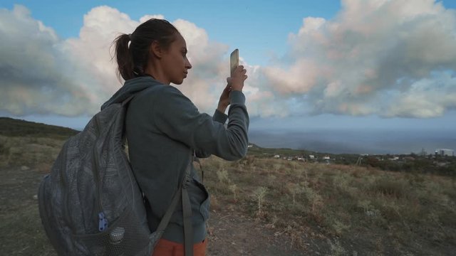 woman hiker looking amazing beautiful cloudy sunset sky and taking photo using mobile phone. female hiker with backpack standind on mountain slope at seaside overlooking of amazing big rose clouds on