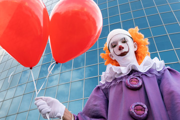 A guy in a scary clown costume with two red balloons against the backdrop of a modern city building. Cosplay for the celebration of Halloween and All Saints Day or at the carnival.