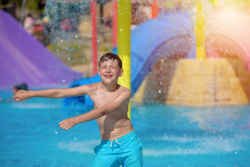 Joyful European boy in blue swimming shorts dancing in water park pool against colorful water slides, he smiling and enjoying his summer holidays.