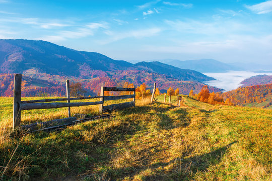 rural area in mountain at sunrise. wonderful golden autumn weather with high clouds on the blue sky. wooden fence along the path through grassy meadow in to the distant valley full of morning fog. nat