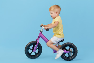 Little child boy learns to keep balance while riding a bicycle. Stylish child in yellow T-shirt on blue background
